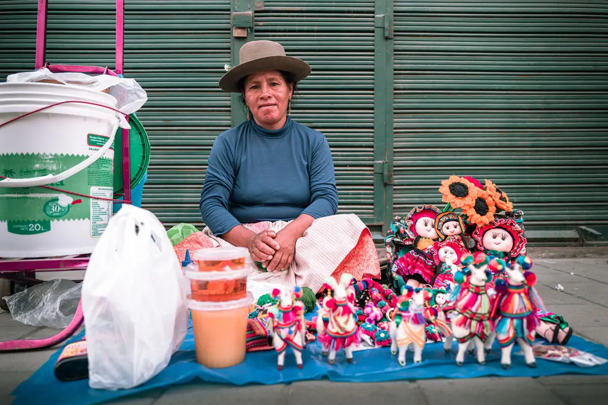 Vendedora andina con sombrero junto a sus muñecas y llamas tejidas en la vereda