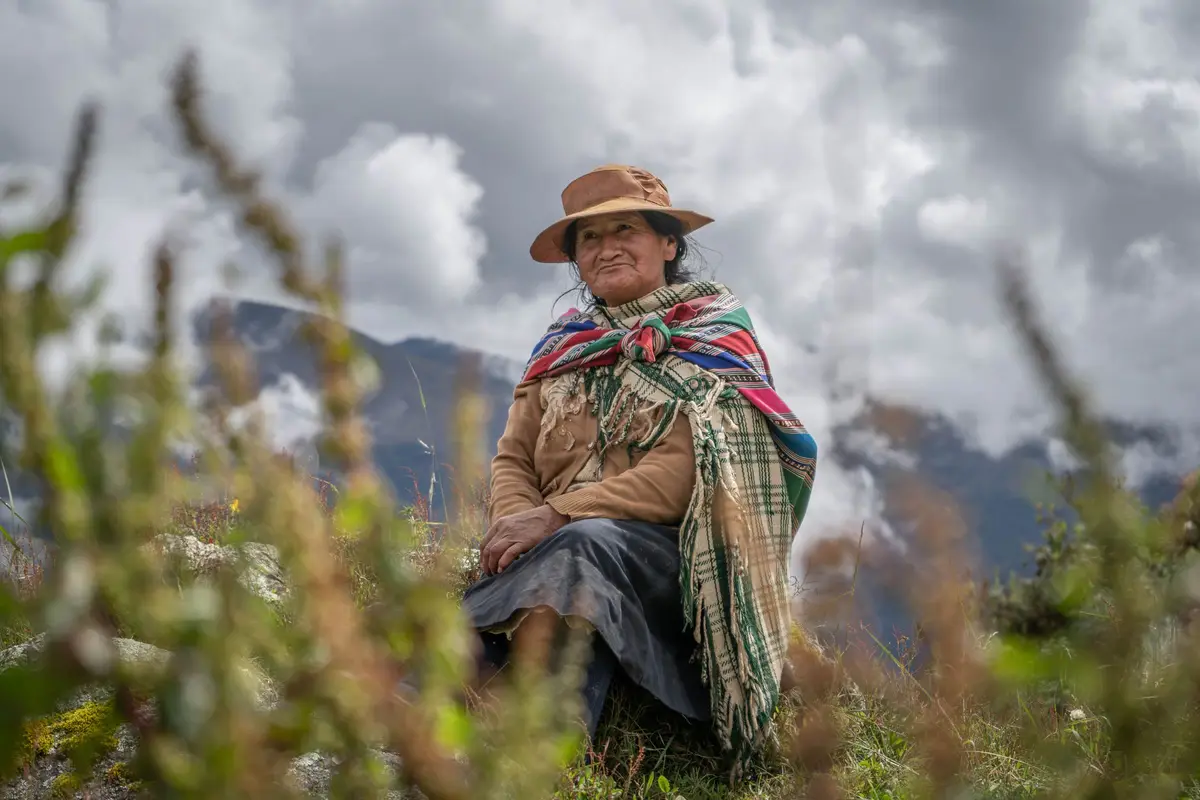 Mujer andina con sombrero y manta a cuadros sentada en el campo frente a los cerros