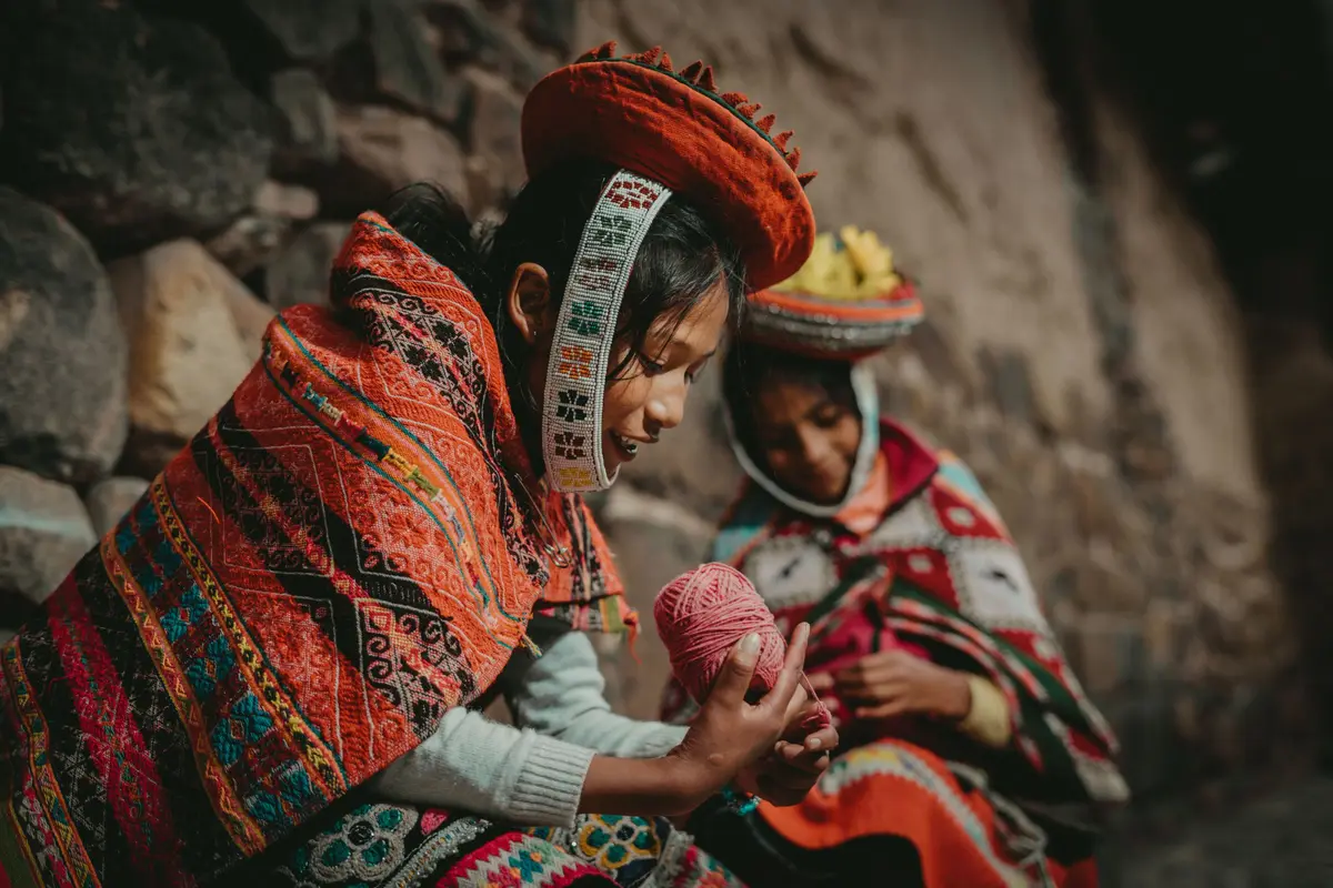 Mujeres con poncho y sombrero tradicional devanando lana junto a un muro de piedra
